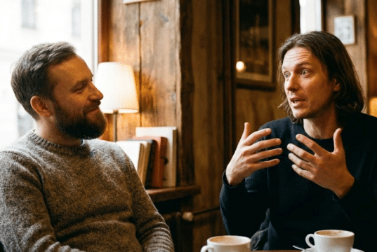 Veidrodžio principas: kaip paprastas atkartojimas padeda užmegzti ryšį? du vyrai kalbasi kavinėje, vienas gestikuliuoja ir pasakoja kitam