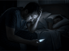 Vienišo vilko mitas: kodėl socialinė izoliacija veda į perdegimą Pavargęs vyras sėdi tamsoje ant lovos ir žiūri į telefoną, simbolizuojantis socialinę izoliaciją ir stresą.