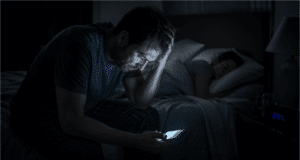 Vienišo vilko mitas: kodėl socialinė izoliacija veda į perdegimą Pavargęs vyras sėdi tamsoje ant lovos ir žiūri į telefoną, simbolizuojantis socialinę izoliaciją ir stresą.
