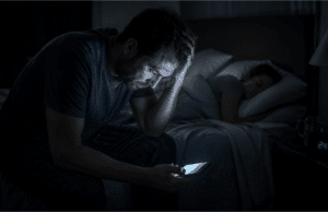 Vienišo vilko mitas: kodėl socialinė izoliacija veda į perdegimą Pavargęs vyras sėdi tamsoje ant lovos ir žiūri į telefoną, simbolizuojantis socialinę izoliaciją ir stresą.