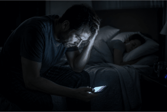 Vienišo vilko mitas: kodėl socialinė izoliacija veda į perdegimą Pavargęs vyras sėdi tamsoje ant lovos ir žiūri į telefoną, simbolizuojantis socialinę izoliaciją ir stresą.