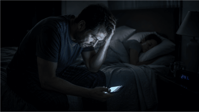 Pavargęs vyras sėdi tamsoje ant lovos ir žiūri į telefoną, simbolizuojantis socialinę izoliaciją ir stresą.